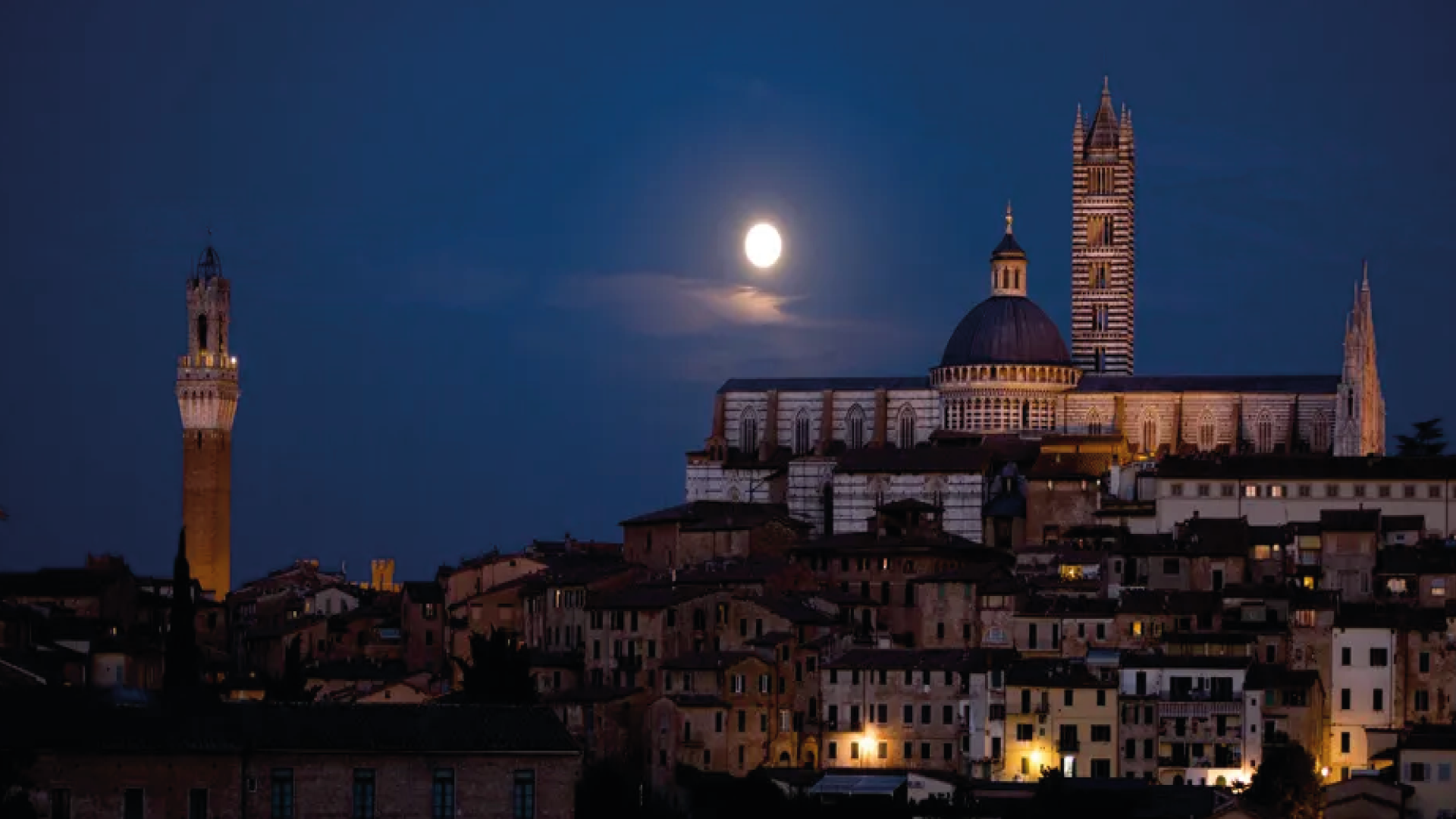 Una cattedrale e un campanile illuminati si ergono su una città storica sotto la luna piena di notte.