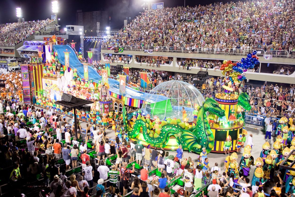 Un char de défilé immense et coloré avec un dragon vert traverse un stade rempli de spectateurs la nuit pendant un carnaval.