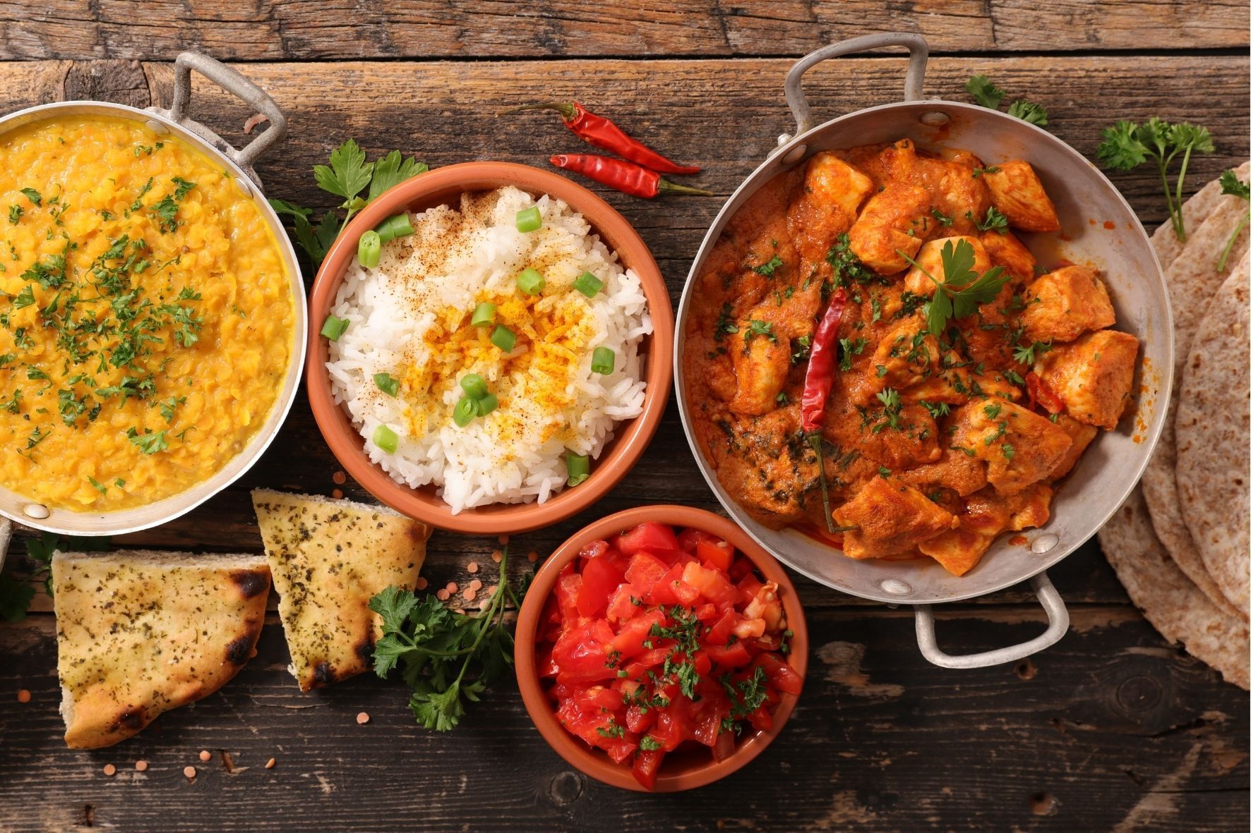 Eine Vogelperspektive auf indische Gerichte, darunter Hühnercurry, Dal, Reis, Salat und Brot, angerichtet auf einem dunklen Holztisch.