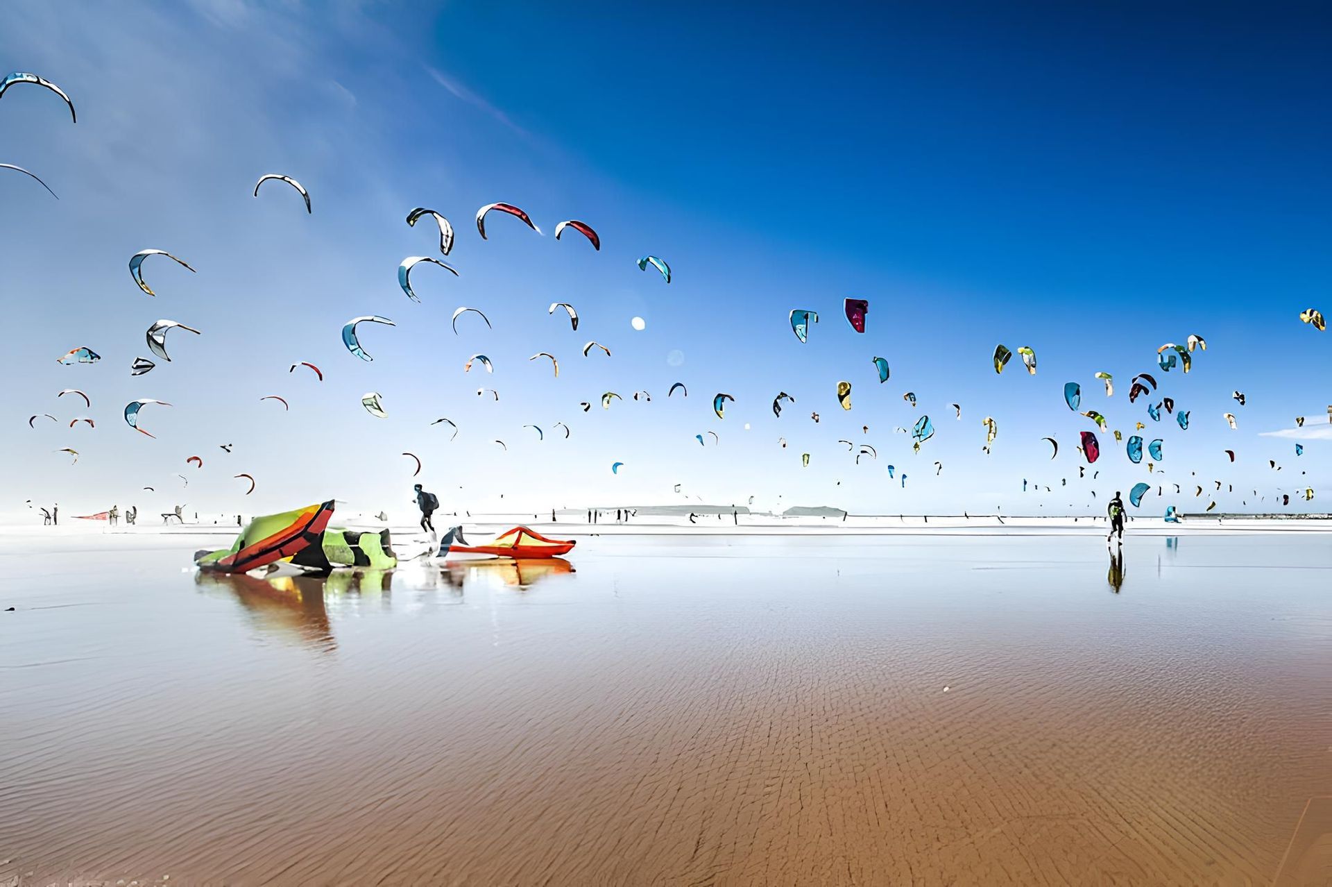 Numerosi aquiloni colorati da kitesurf riempiono un cielo azzurro limpido sopra una spiaggia bagnata e riflettente con persone.