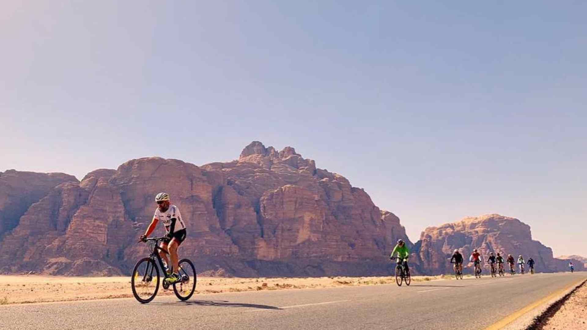 Un viaggio di gruppo WeRoad con persone in bicicletta su una strada asfaltata attraverso un paesaggio desertico con grandi formazioni rocciose sullo sfondo.