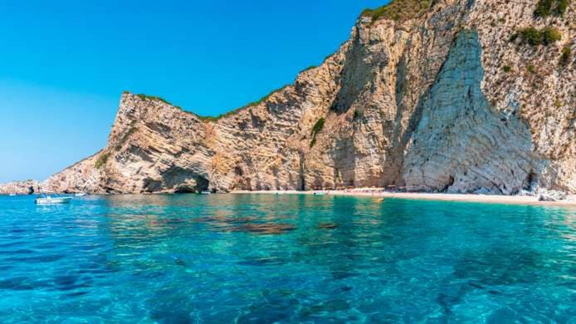 Una grande scogliera rocciosa si erge su una spiaggia sabbiosa isolata e un mare limpido e turchese sotto un cielo blu.