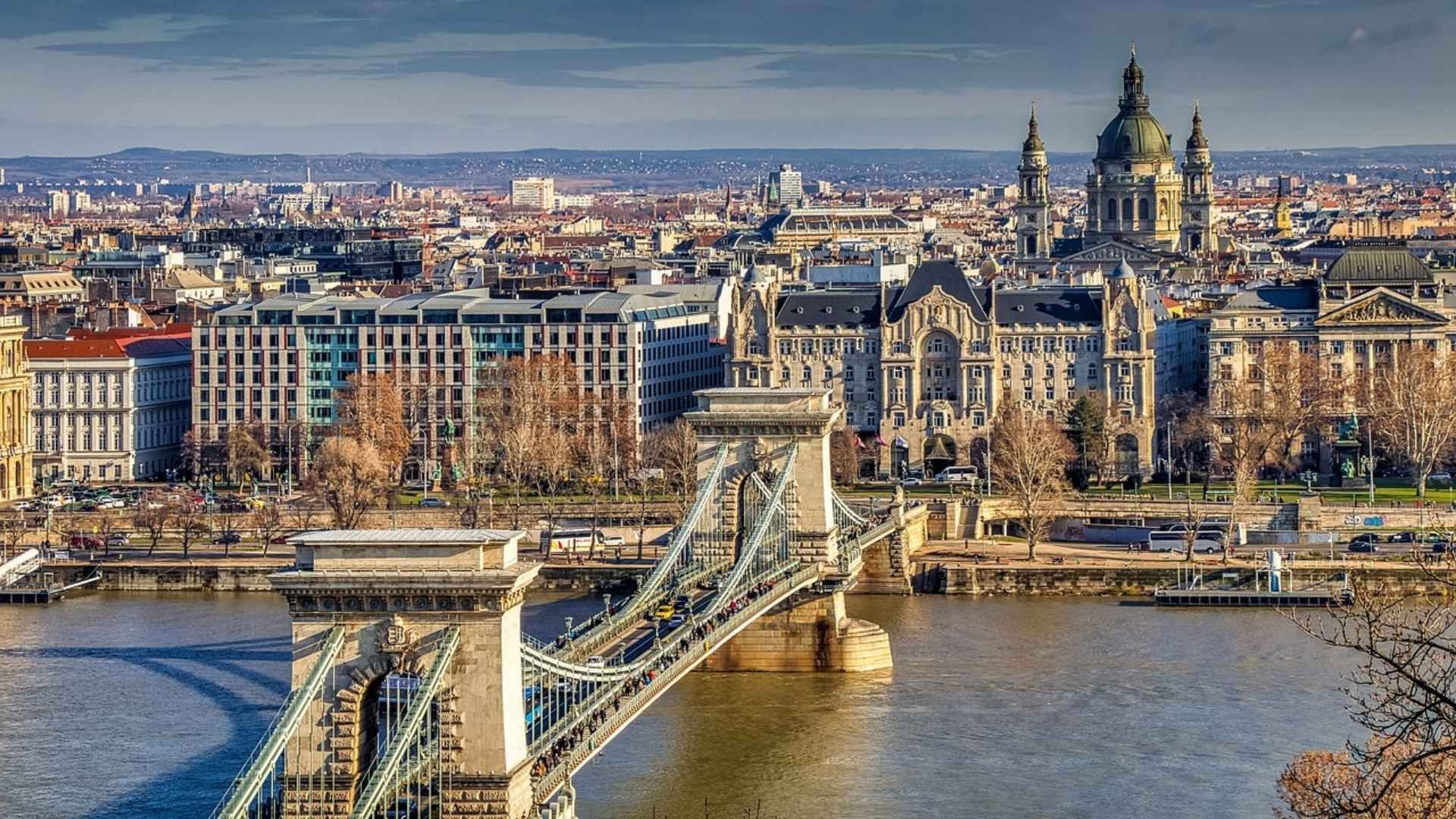 Un grand pont suspendu enjambe une rivière devant un paysage urbain dense, comprenant des bâtiments historiques et une cathédrale à dôme.