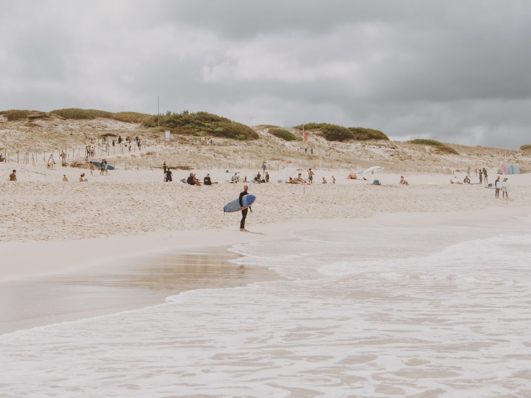 Un surfista con traje de neopreno sostiene una tabla de surf azul a la orilla del agua en una playa concurrida con dunas de arena bajo un cielo nublado.