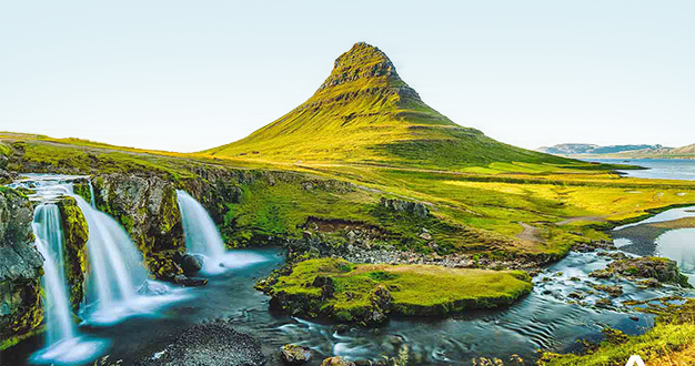A wide-angle shot of a green, conical mountain with waterfalls cascading into a river in the foreground, under a clear sky.