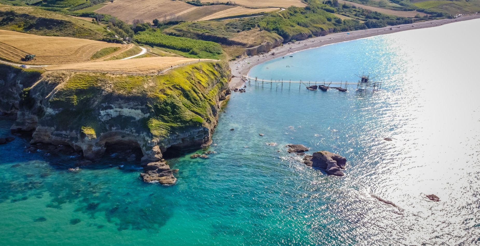 Veduta aerea di un pontile in legno che si protende da una costa rocciosa e rigogliosa verso un mare turchese, con dolci colline alle spalle.