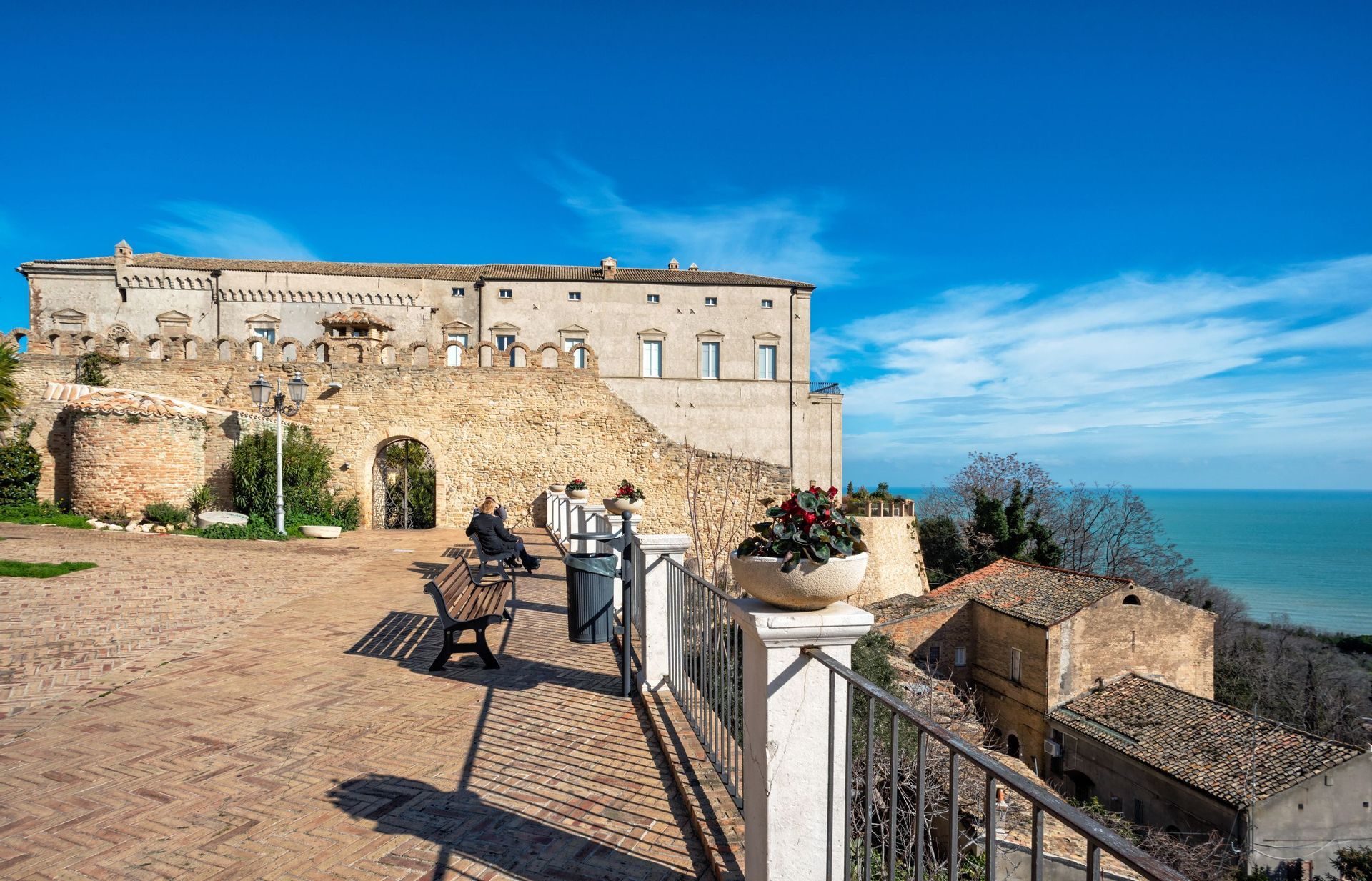 Un'ampia terrazza in mattoni con panchine si trova di fronte a un grande edificio in pietra, affacciata sul mare azzurro brillante sotto un cielo sereno.
