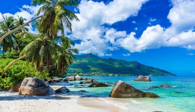 Palme rigogliose su una spiaggia di sabbia bianca con grandi massi, bagnata da acque cristalline turchesi, di fronte a una catena montuosa verde.