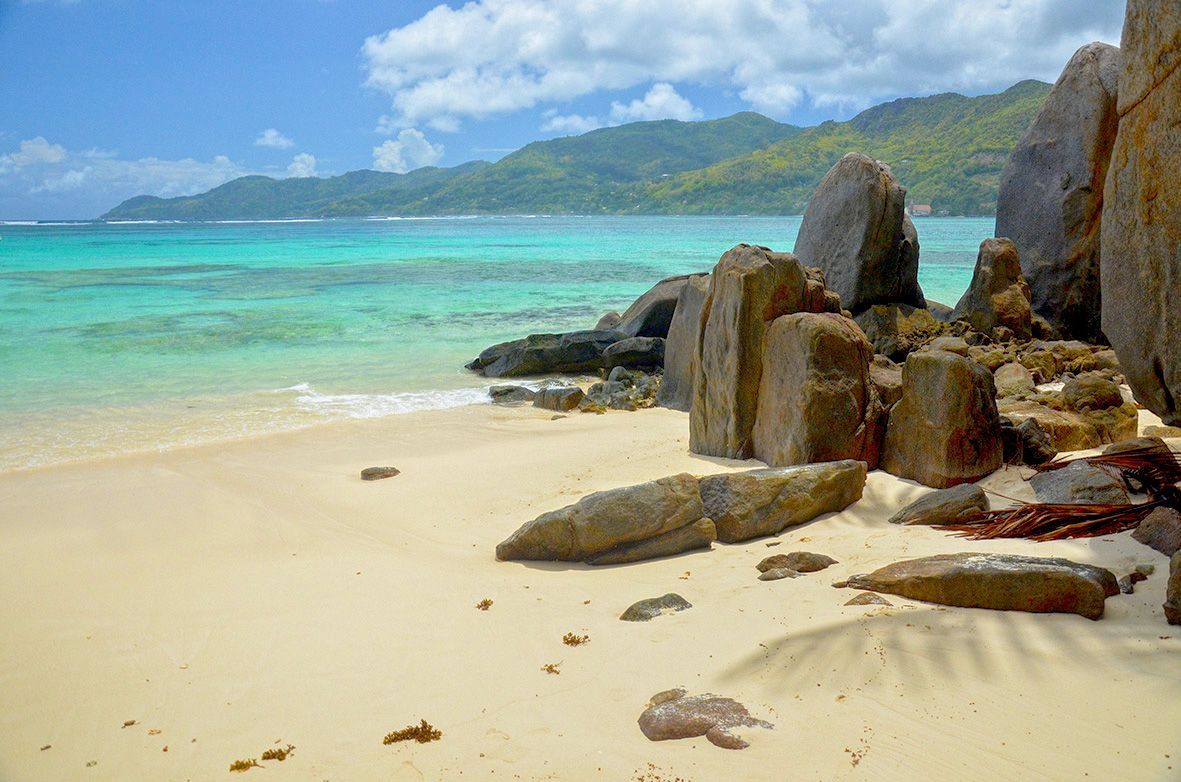 Grandi formazioni rocciose si ergono su una spiaggia sabbiosa accanto ad acque turchesi, con verdi colline visibili in lontananza sotto un cielo blu.
