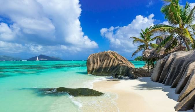 Grossi massi si trovano su una spiaggia di sabbia bianca accanto ad acque turchesi, con palme sotto un cielo parzialmente nuvoloso.