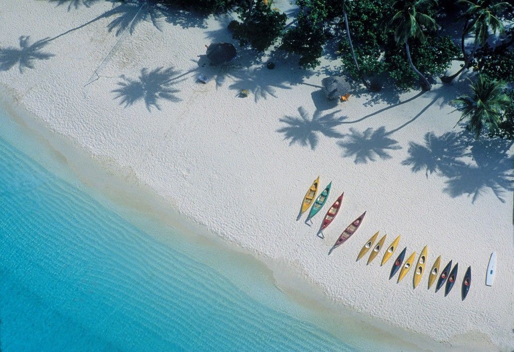 Una vista aerea di kayak colorati allineati su una spiaggia di sabbia bianca accanto all'acqua turchese, con le ombre delle palme.