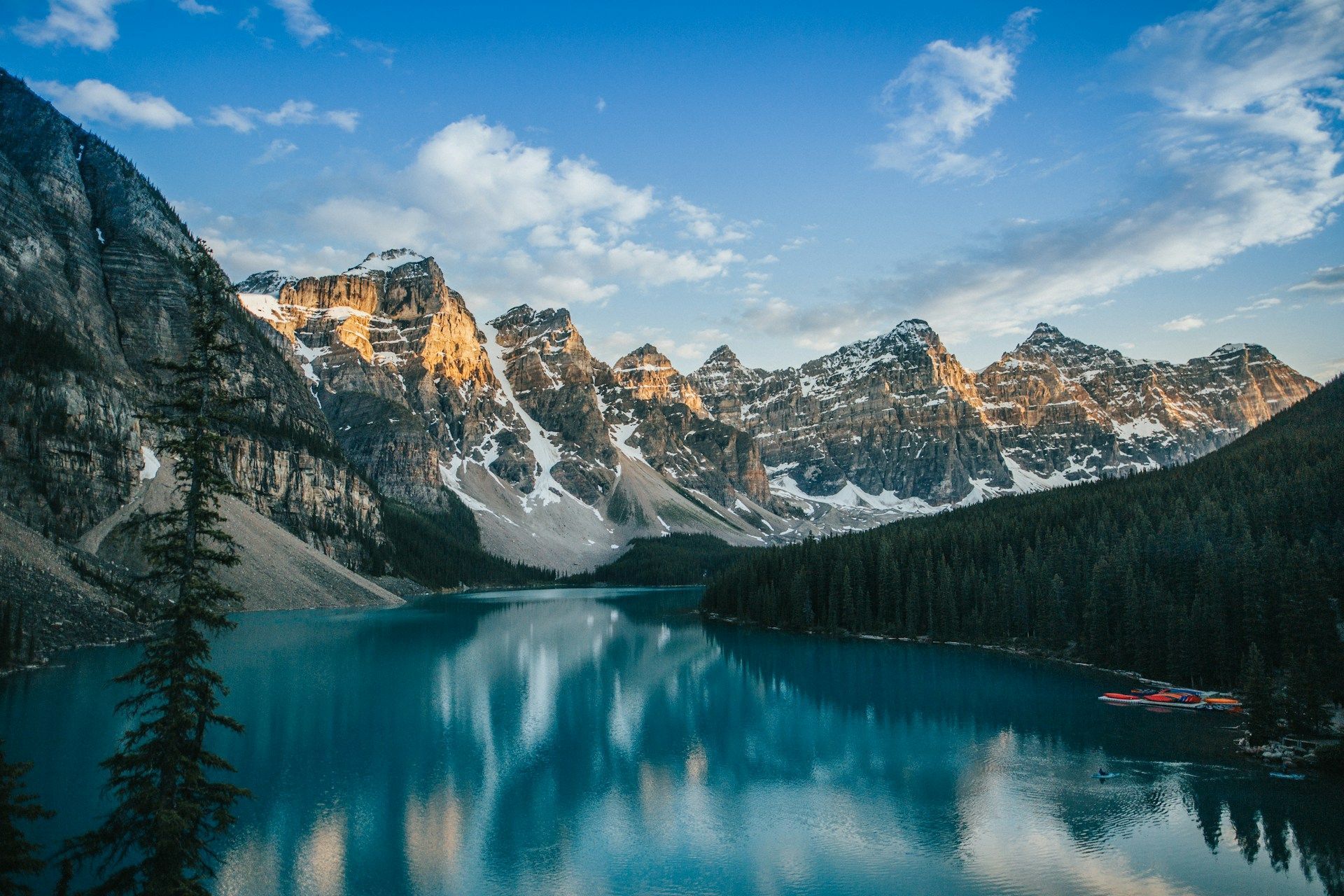 Sunlight hits the peaks of snow-capped mountains which reflect in a calm, turquoise lake surrounded by a pine forest.