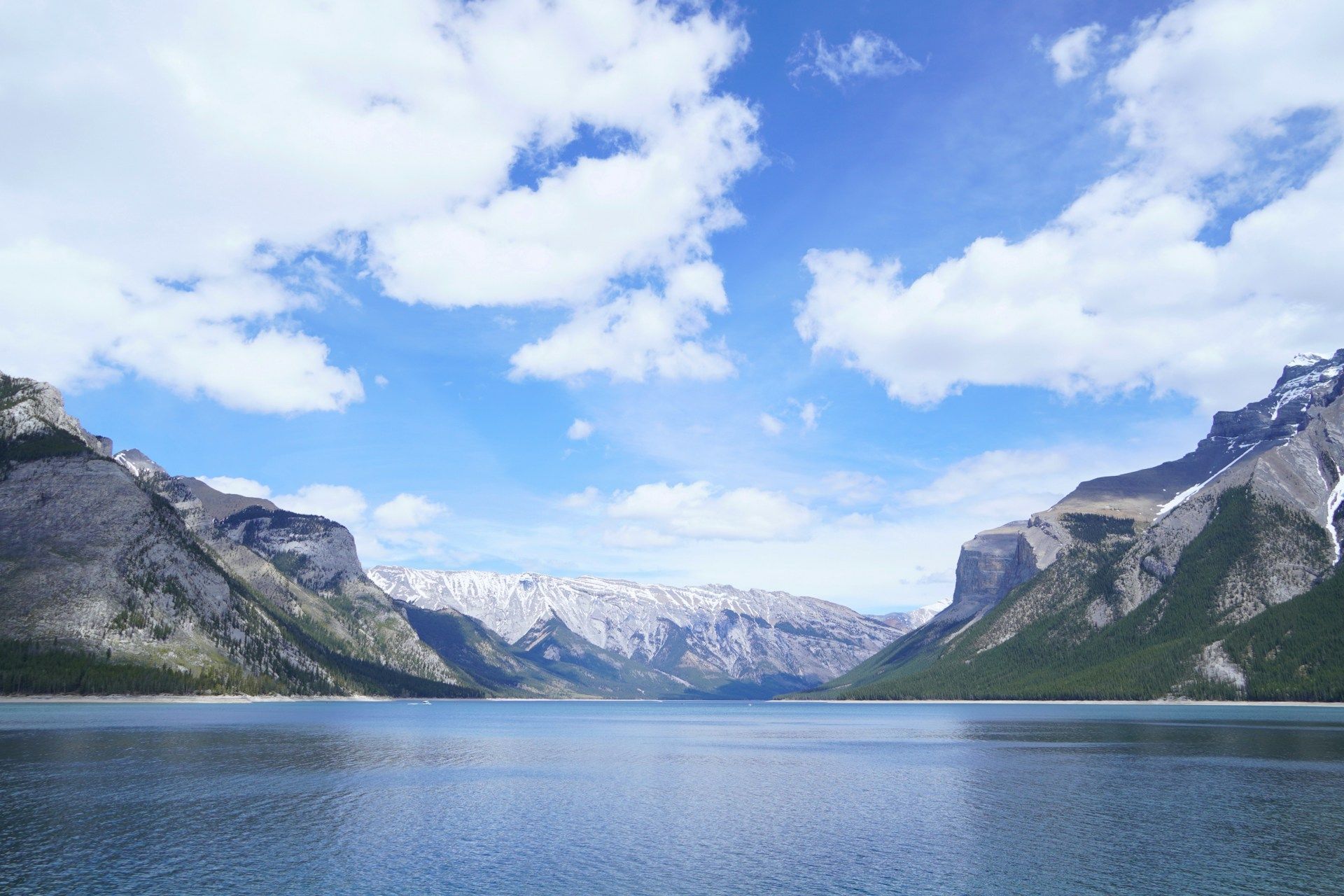 A wide, calm lake is flanked by steep, forested mountains under a blue sky with fluffy white clouds.
