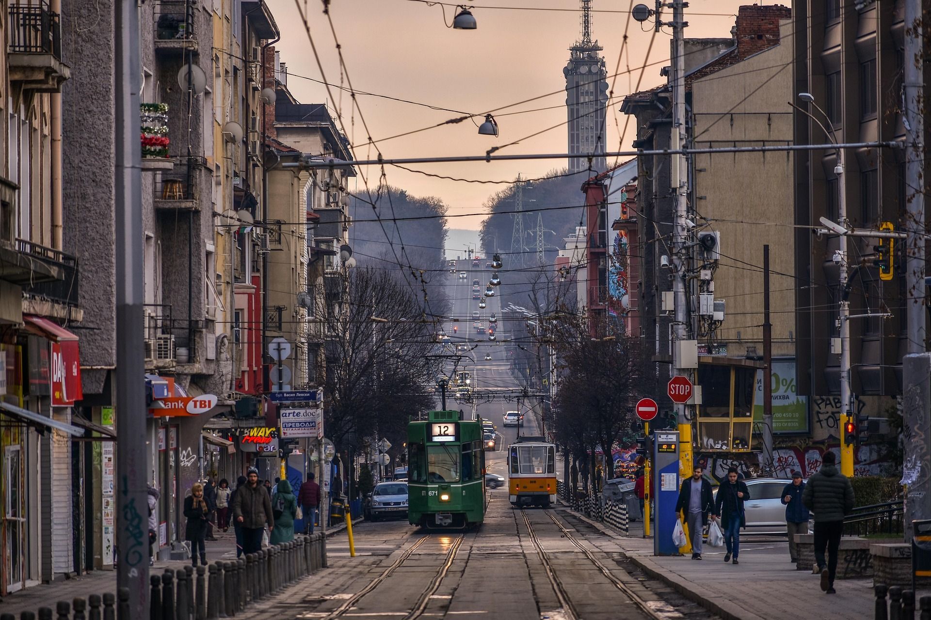 Veduta lungo una lunga via cittadina con tram sui binari, pedoni sui marciapiedi e una torre delle telecomunicazioni in lontananza.