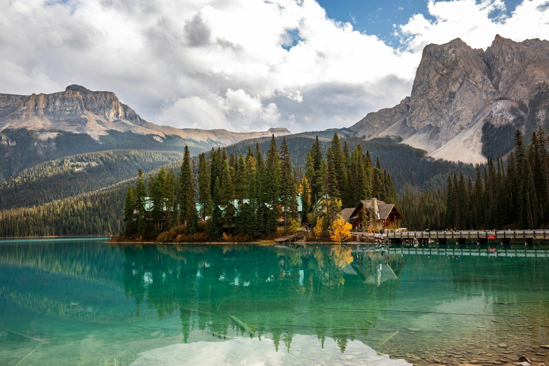 A wooden lodge on a forested peninsula is reflected in a turquoise alpine lake with mountains in the background.