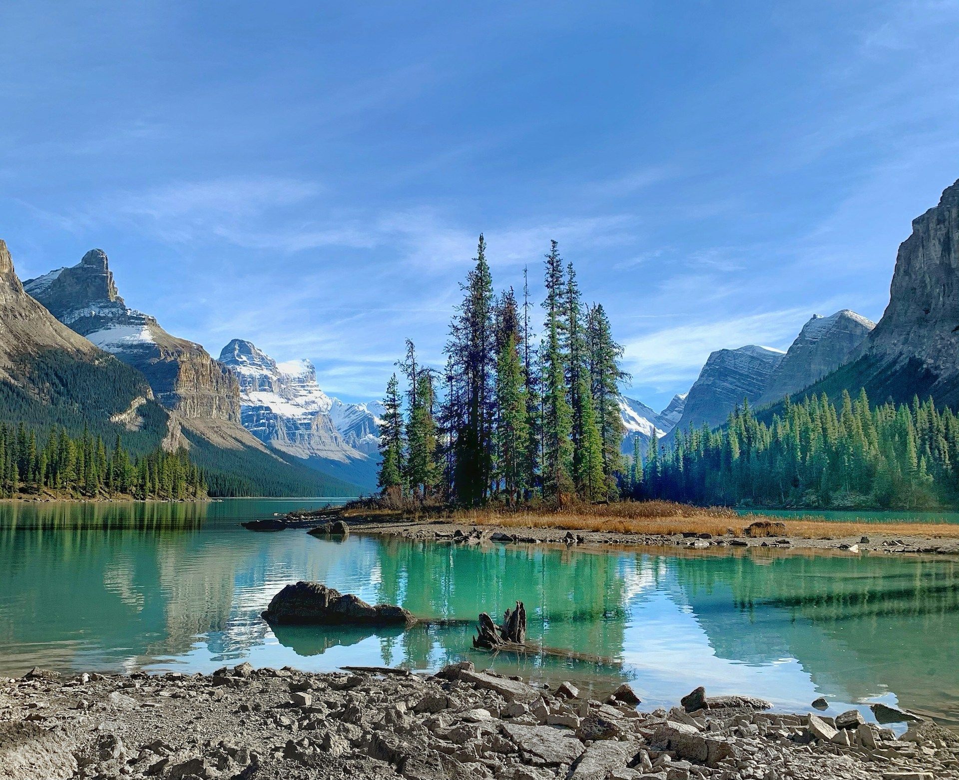 A small island with pine trees sits in a calm turquoise lake, surrounded by forests and snow-capped mountains.