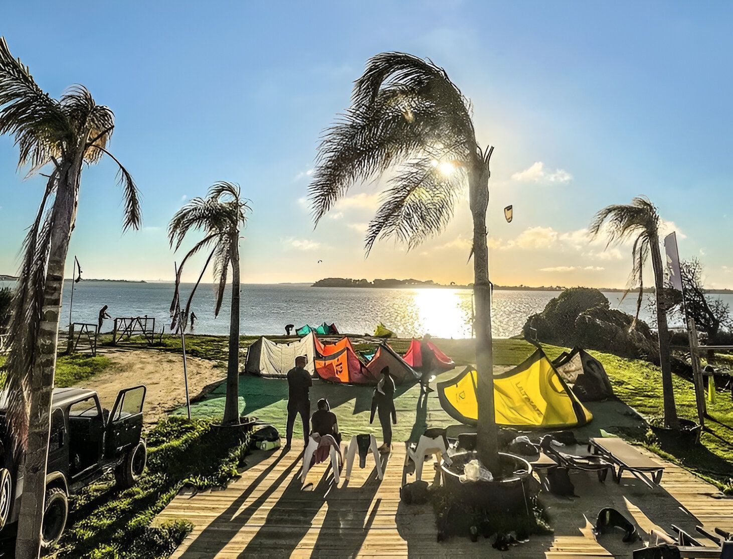 Un viaggio di gruppo WeRoad si prepara per il kitesurf su una spiaggia con palme mentre il sole tramonta sull'acqua.