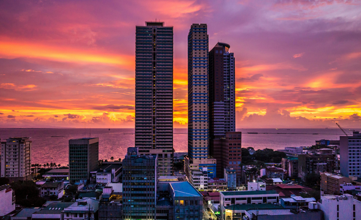 Hohe Wolkenkratzer überblicken eine Küstenstadt unter einem dramatischen pink-orangen Sonnenuntergangshimmel.