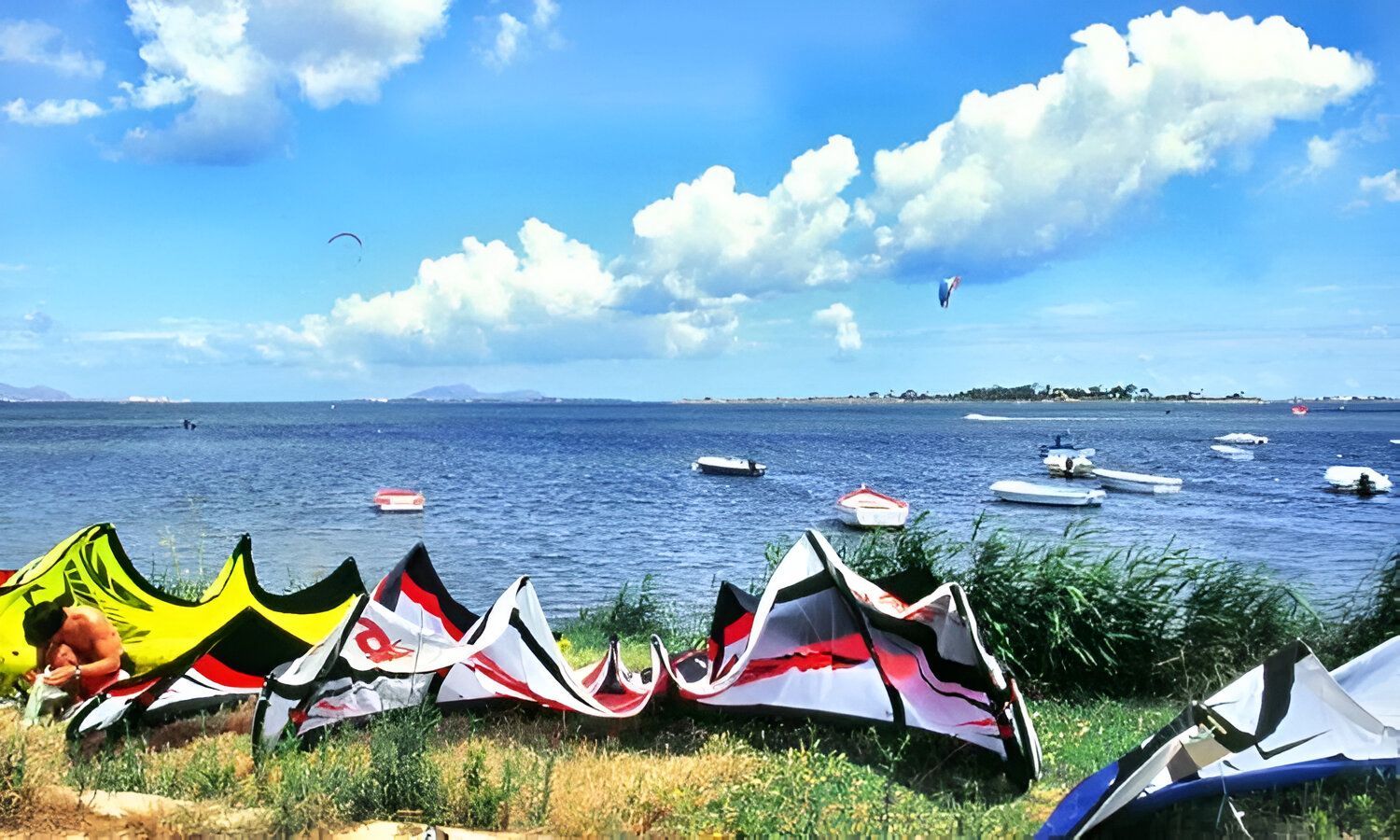 Kitesurf colorati riposano su una sponda erbosa in riva al mare, con barche ormeggiate nell'acqua sotto un cielo nuvoloso.