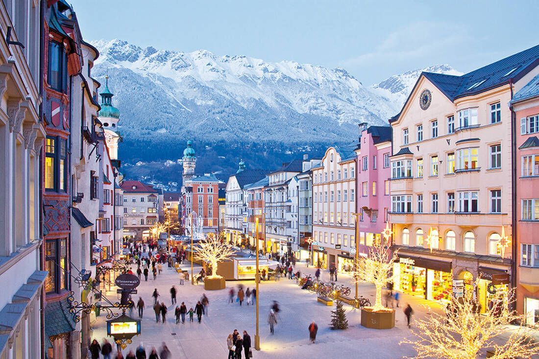 Una vivace via pedonale affollata, fiancheggiata da edifici colorati e alberi illuminati, con uno sfondo di montagne innevate.
