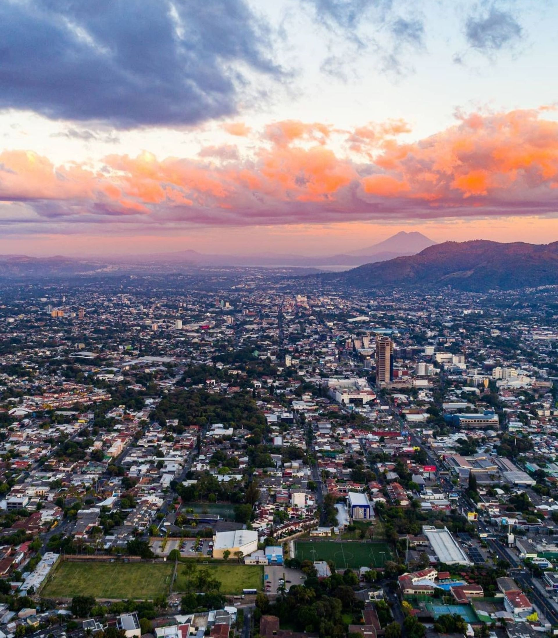 Eine Luftaufnahme einer weitläufigen Stadtlandschaft bei Sonnenuntergang, mit orangefarbenen Wolken darüber und Bergen am Horizont.