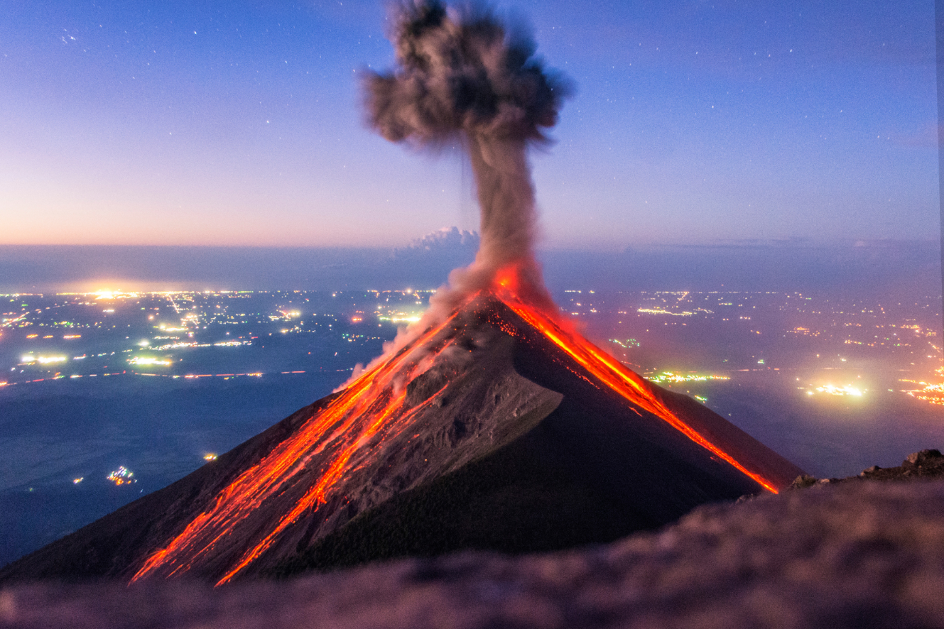 Ein nachts ausbrechender Vulkan, mit Flüssen aus geschmolzener Lava, die seine Hänge hinabfließen, und Rauch, der in einen sternenklaren Himmel über einer Stadt aufsteigt.
