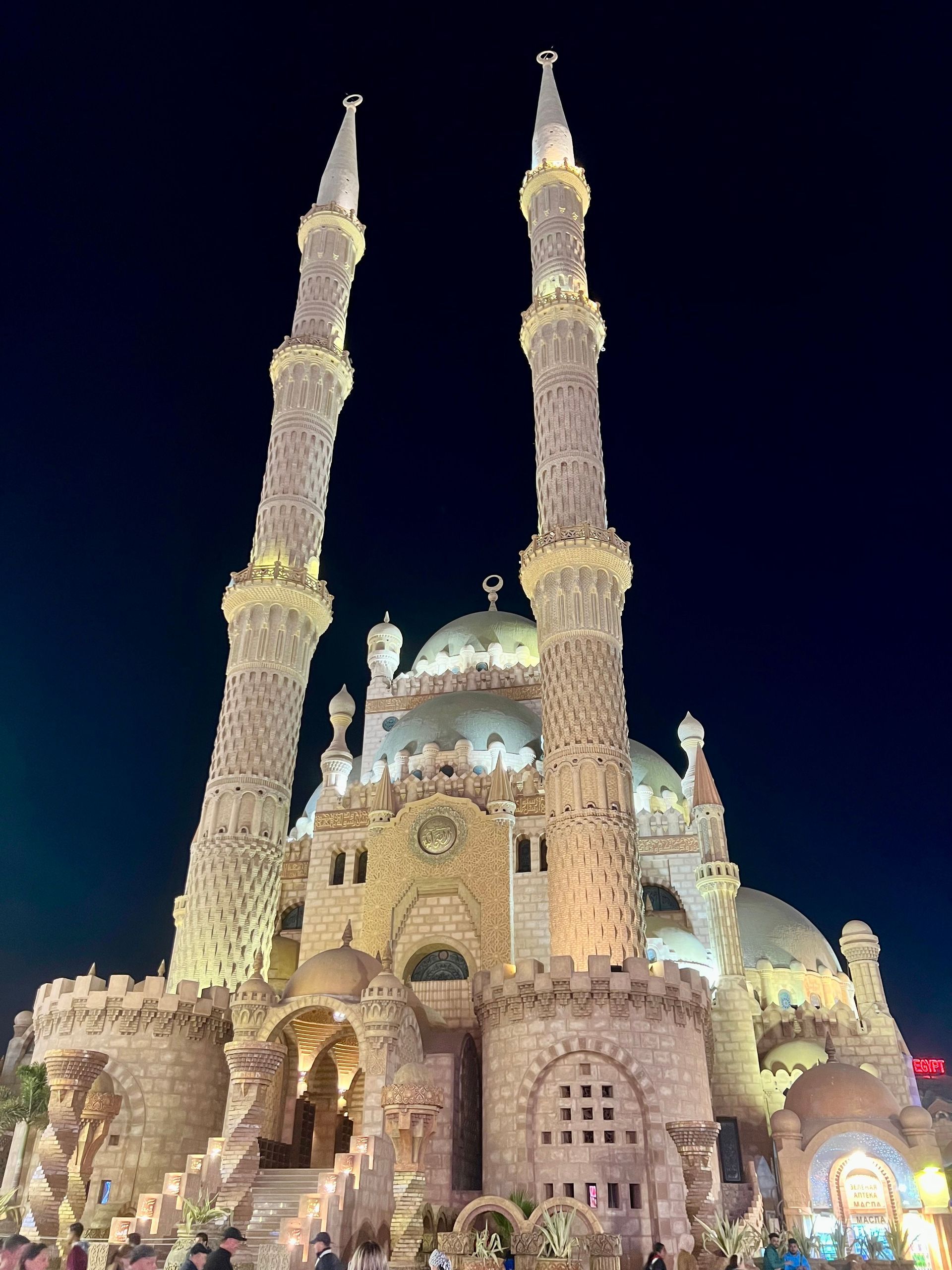 Una moschea in pietra ornata con grandi cupole e due alti minareti è illuminata da luci contro un cielo notturno scuro.