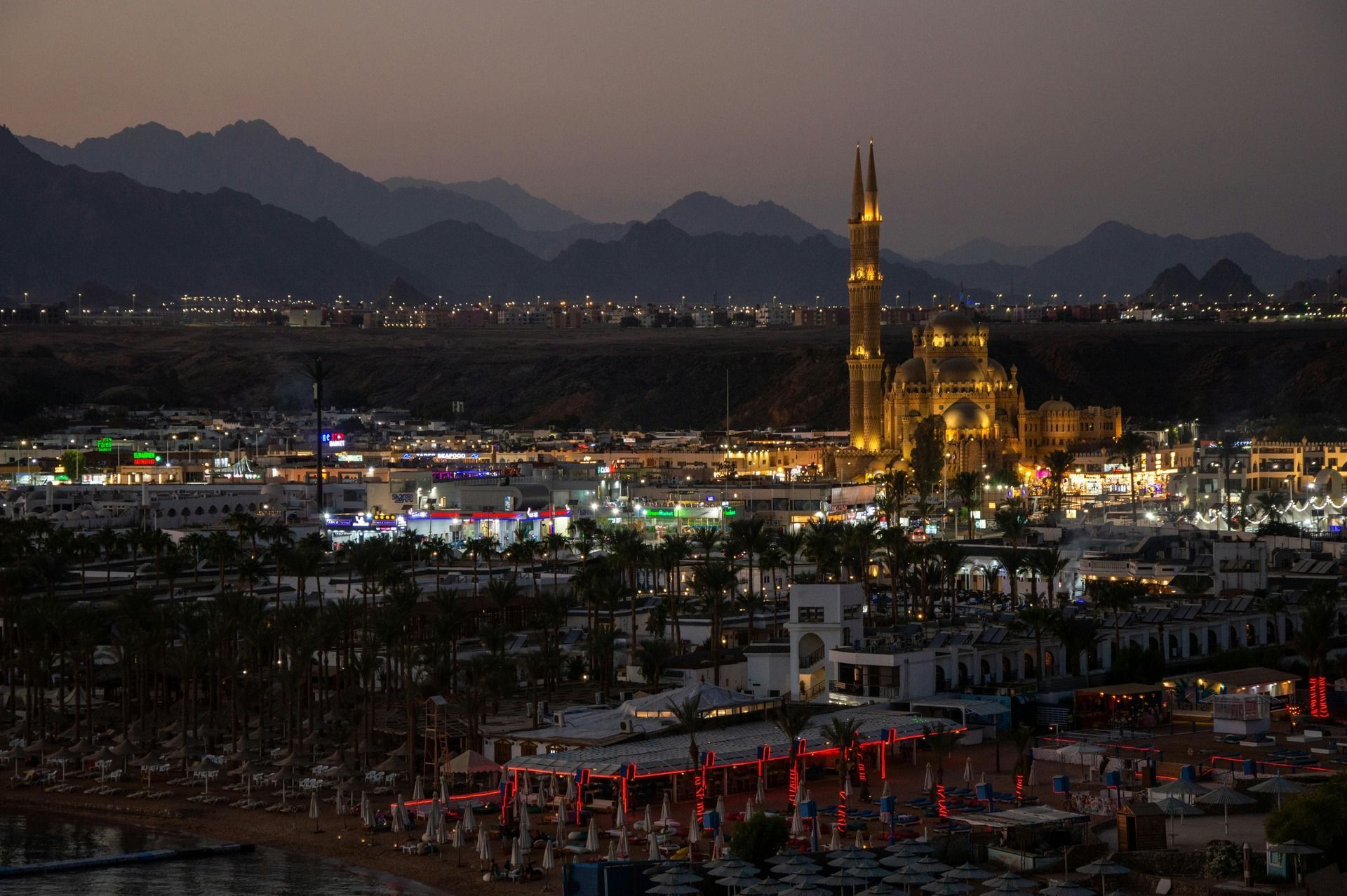 Una vista panoramica di una città costiera al tramonto, con una moschea illuminata e montagne sullo sfondo.