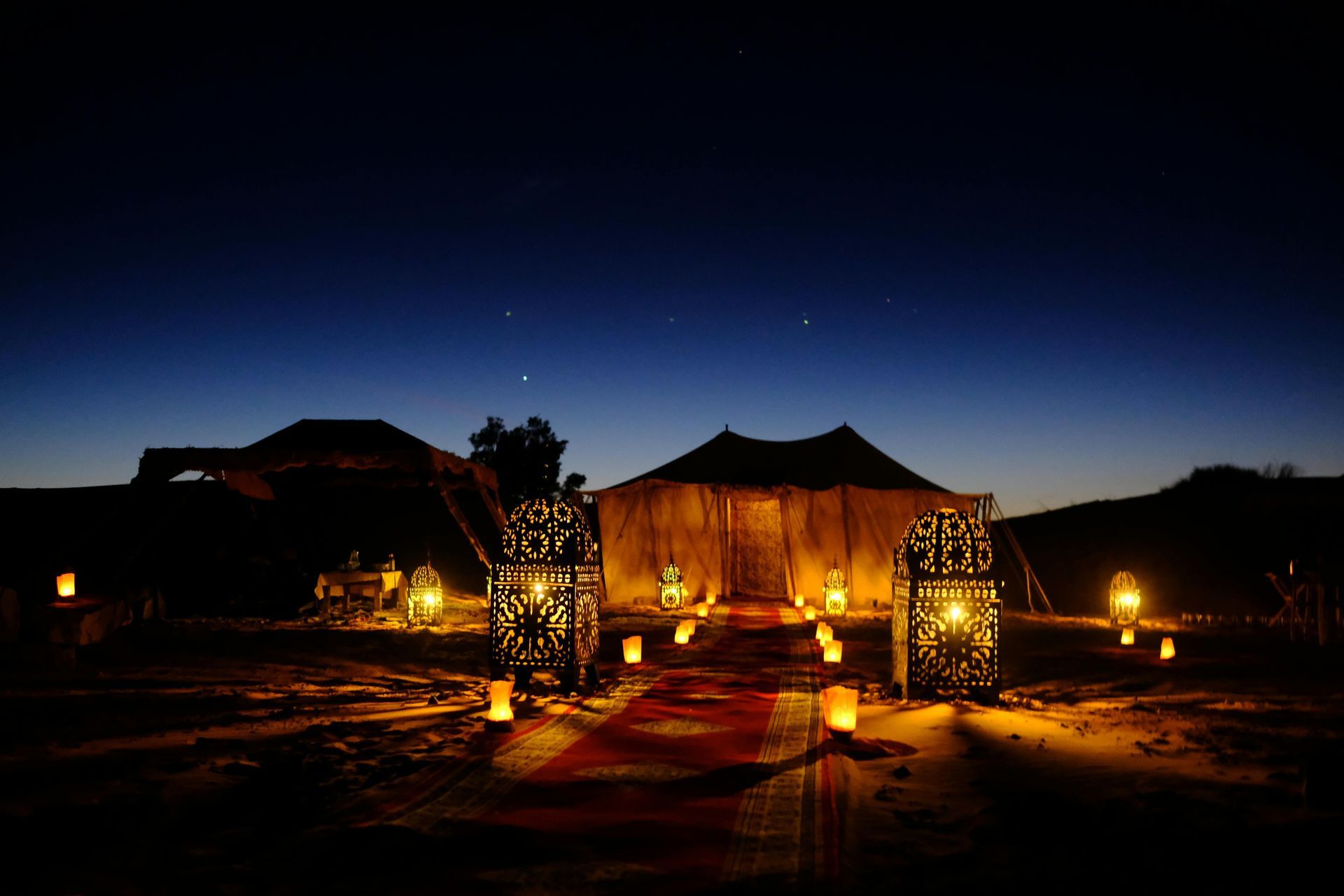 Lanterne illuminate e candele fiancheggiano un sentiero di tappeto rosso verso una tenda nel deserto sotto un cielo notturno stellato.