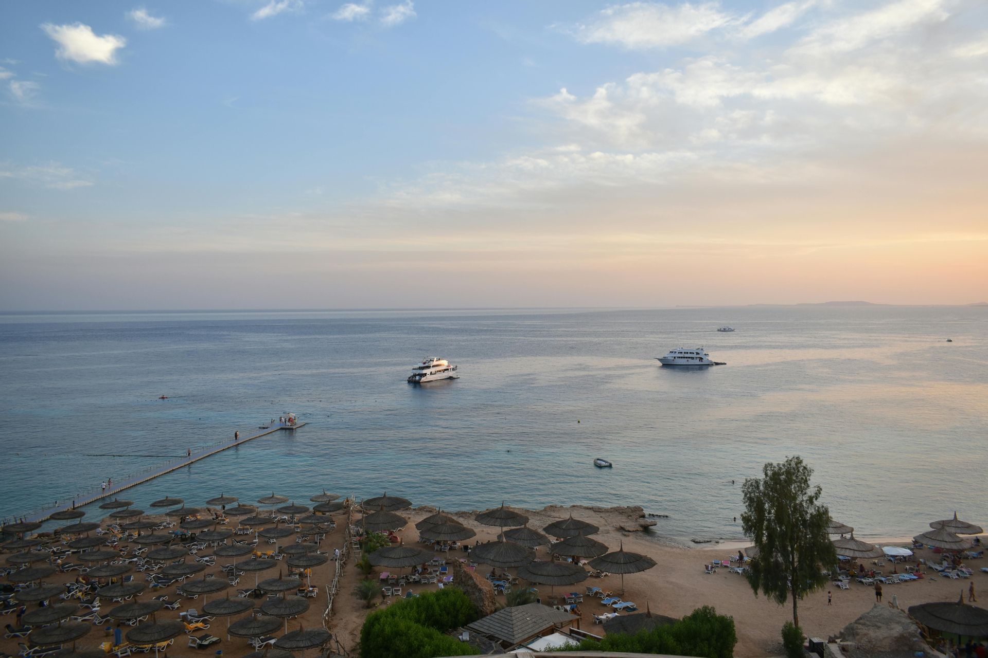Una vista dall'alto di una spiaggia sabbiosa con ombrelloni di paglia e sedie a sdraio, con diversi yacht sul mare calmo al tramonto.