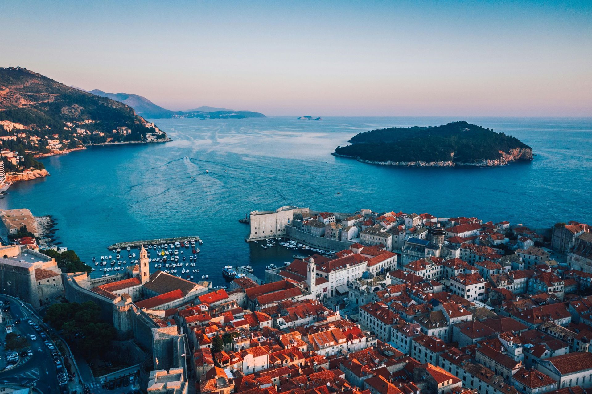 Una vista aerea di una storica città costiera con tetti in tegole rosse, un porto con barche e un'isola nel mare blu all'alba.