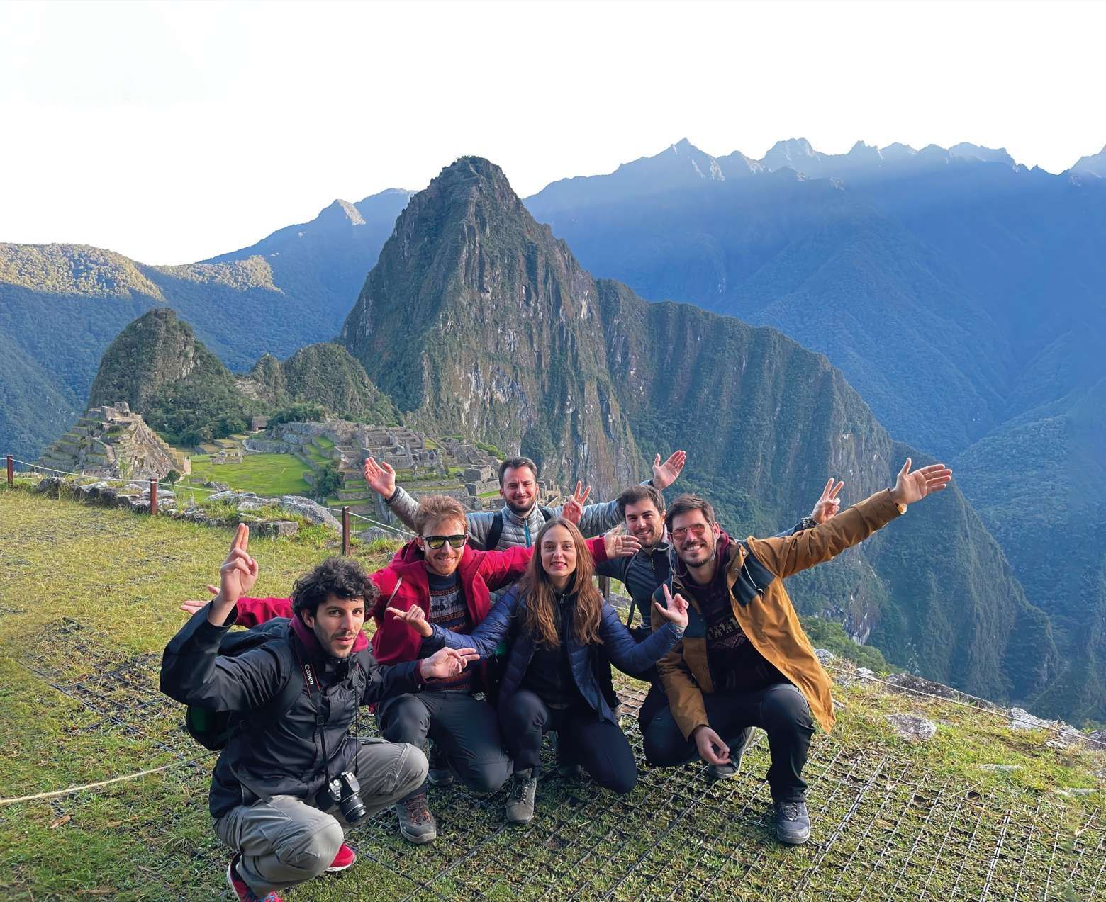 A WeRoad group trip posing for a photo on a grassy viewpoint with ancient ruins and steep green mountains in the background.