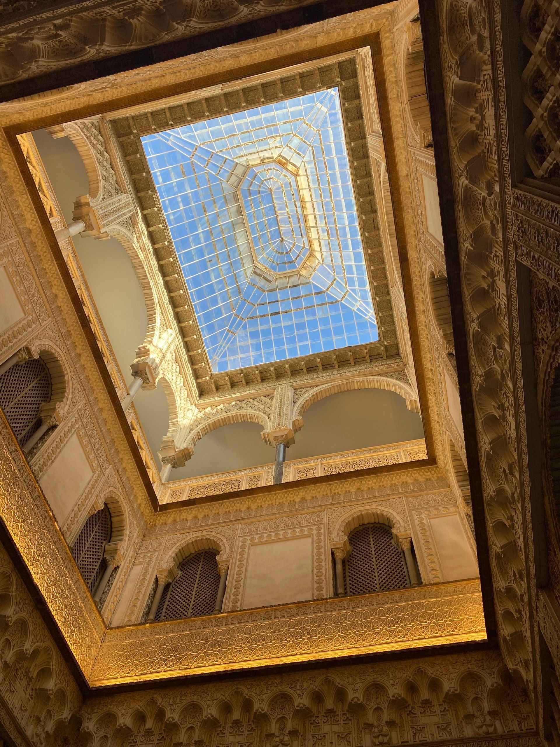 Una vista dal basso attraverso un cortile ornato con archi scolpiti, verso un soffitto di vetro che rivela un cielo blu.