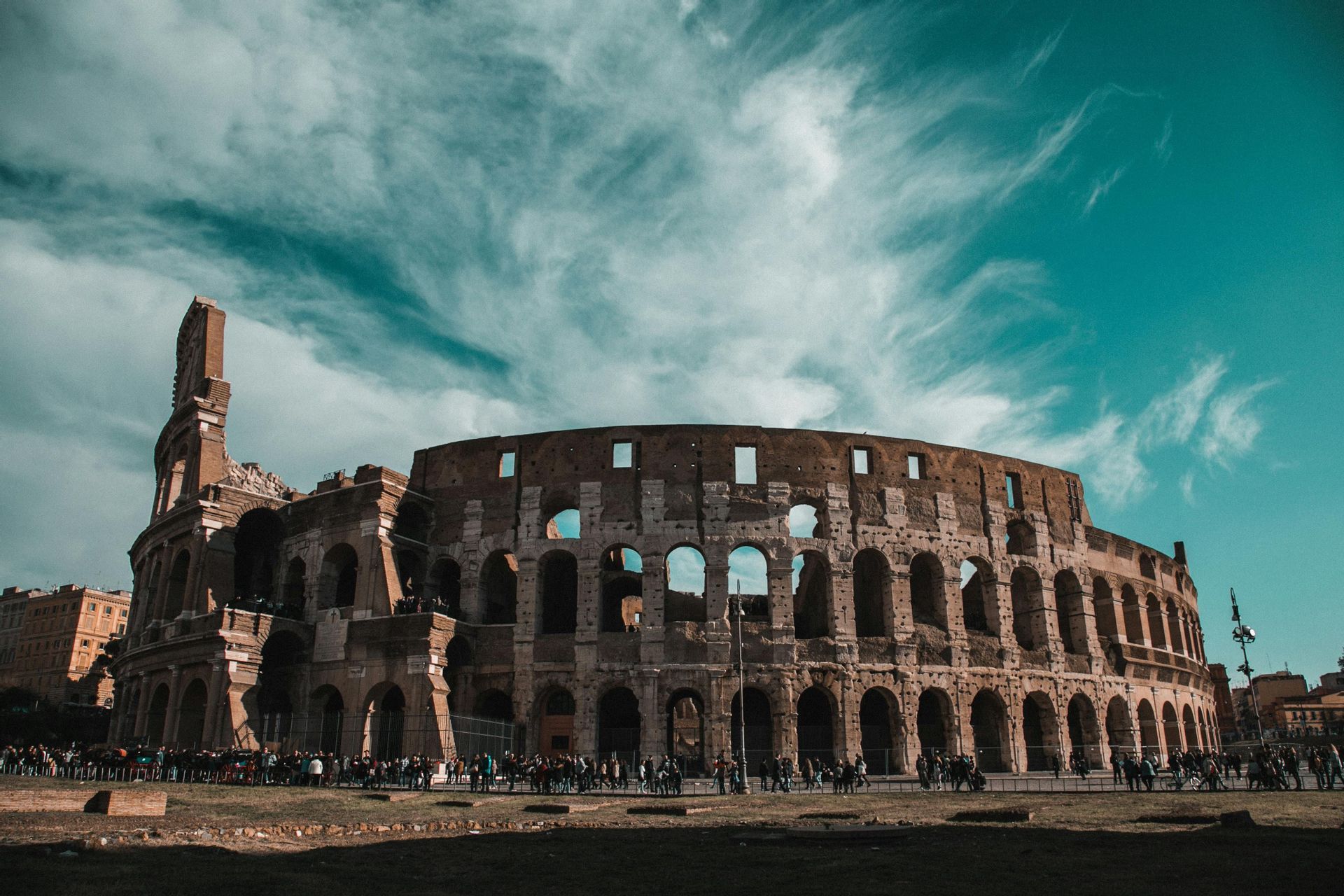 Un grande e antico anfiteatro in pietra si erge sotto un cielo turchese pieno di nuvole leggere, con una folla di persone radunata davanti.