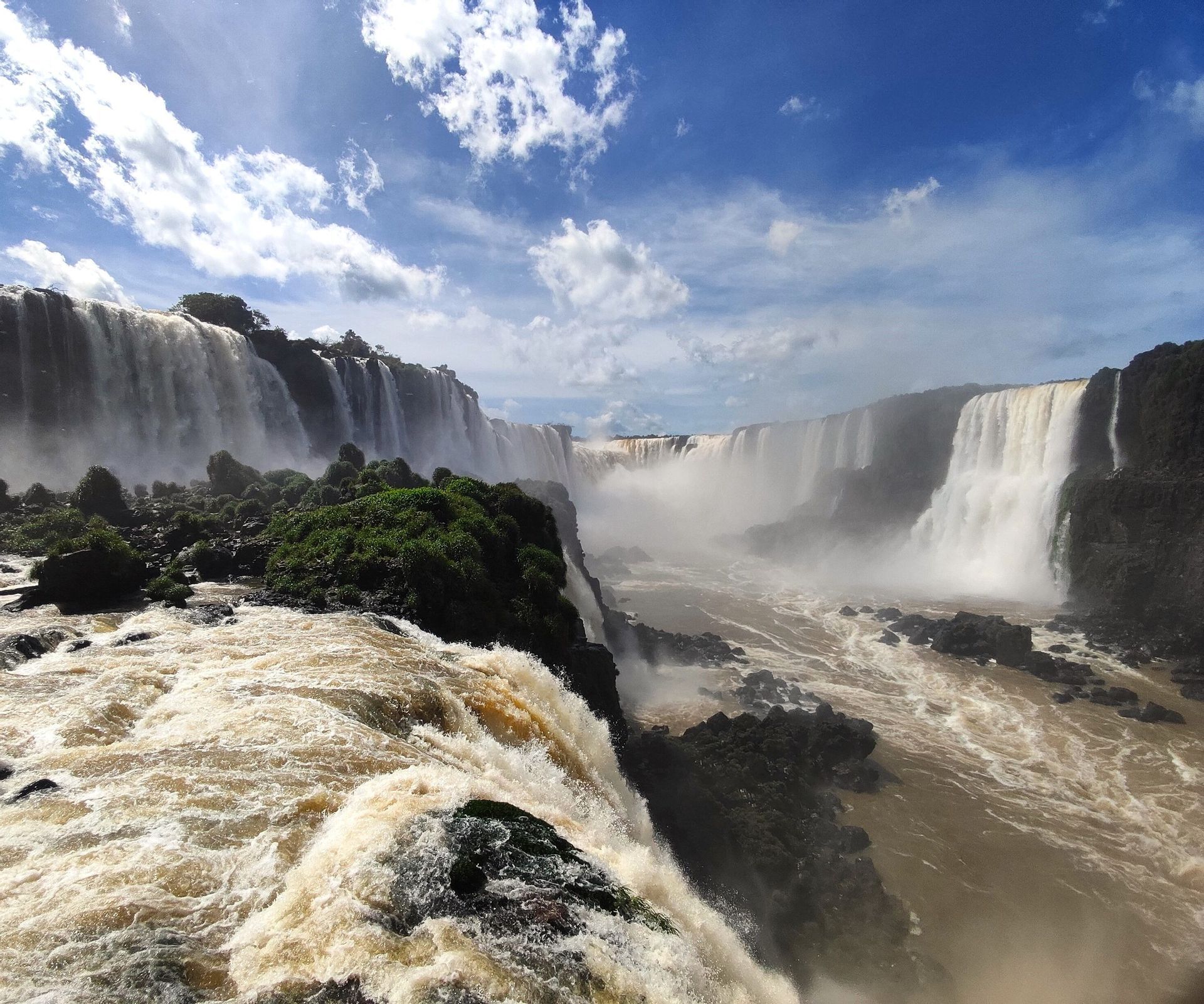 Inmensas cascadas caen sobre acantilados rocosos y exuberantes hacia un cañón neblinoso bajo un cielo azul con nubes blancas.