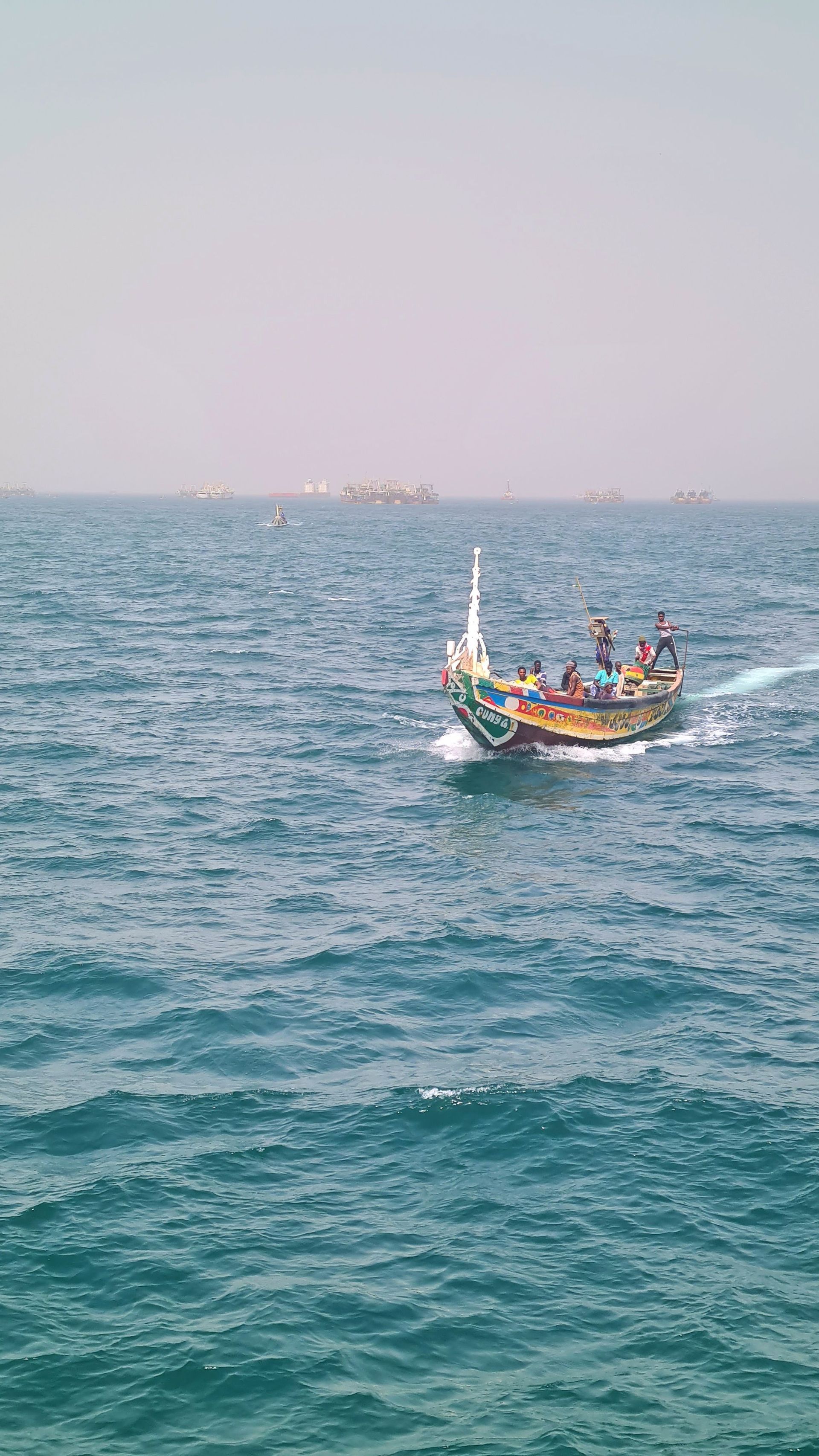Eine Gruppe von Menschen auf einem farbenfrohen Holzboot segelt über ein unruhiges blaues Meer, mit weiteren Booten in der diesigen Ferne.