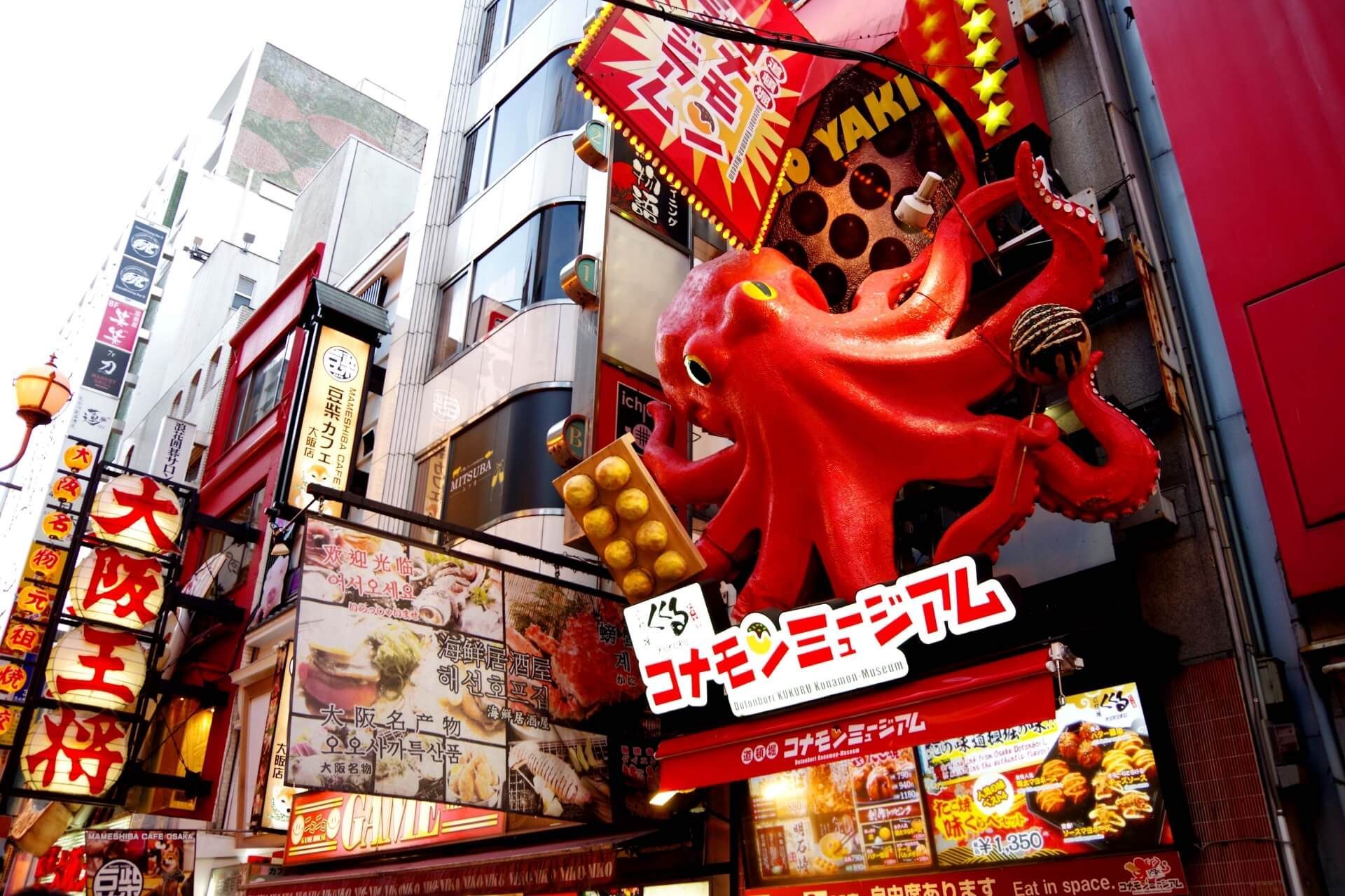 Una scultura gigante di polpo rosso che tiene takoyaki è montata su un edificio sopra una trafficata strada giapponese con molte insegne colorate.