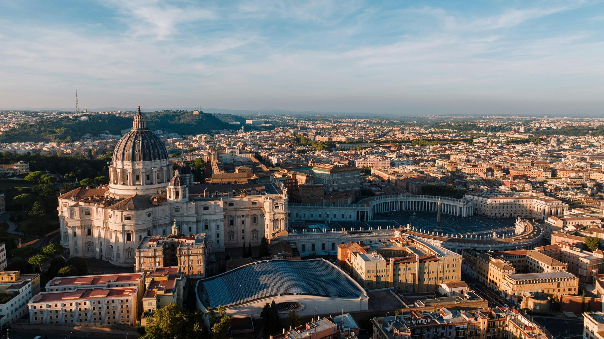 Una vista aerea di una grande basilica con una cupola imponente e una vasta piazza colonnata, che si affaccia su una città estesa all'alba.