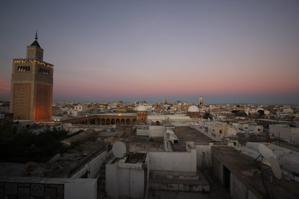 Un minaret illuminé domine un paysage urbain de bâtiments blancs et de toits plats sous un ciel rose et violet au crépuscule.