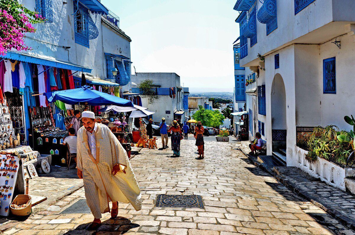 Un homme en tenue traditionnelle marche dans une rue pavée animée bordée d'étals de marché et de bâtiments blanchis à la chaux avec des garnitures bleues.
