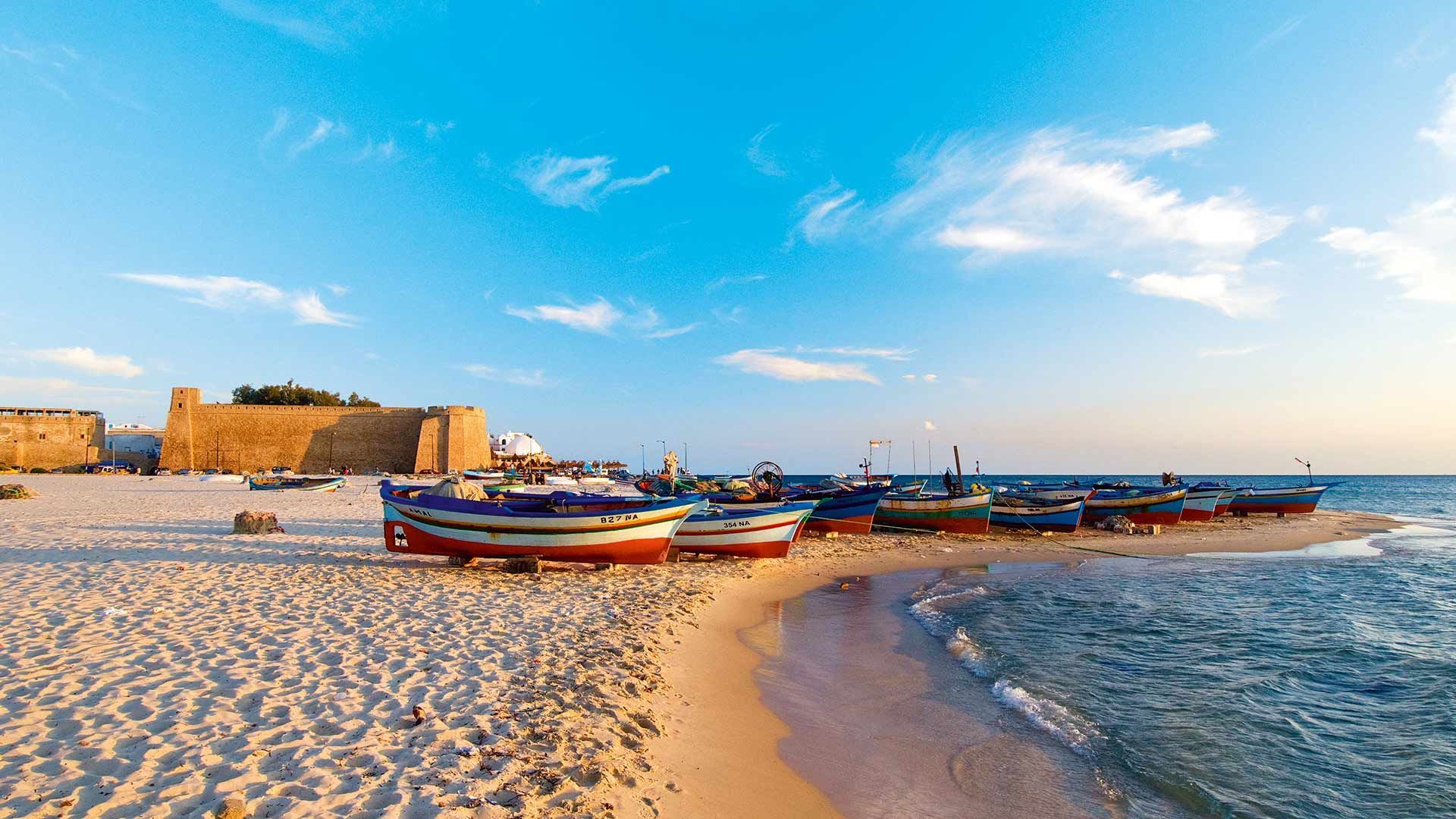 Des bateaux de pêche colorés sont alignés sur une plage de sable près de la mer, avec une grande forteresse en pierre en arrière-plan.