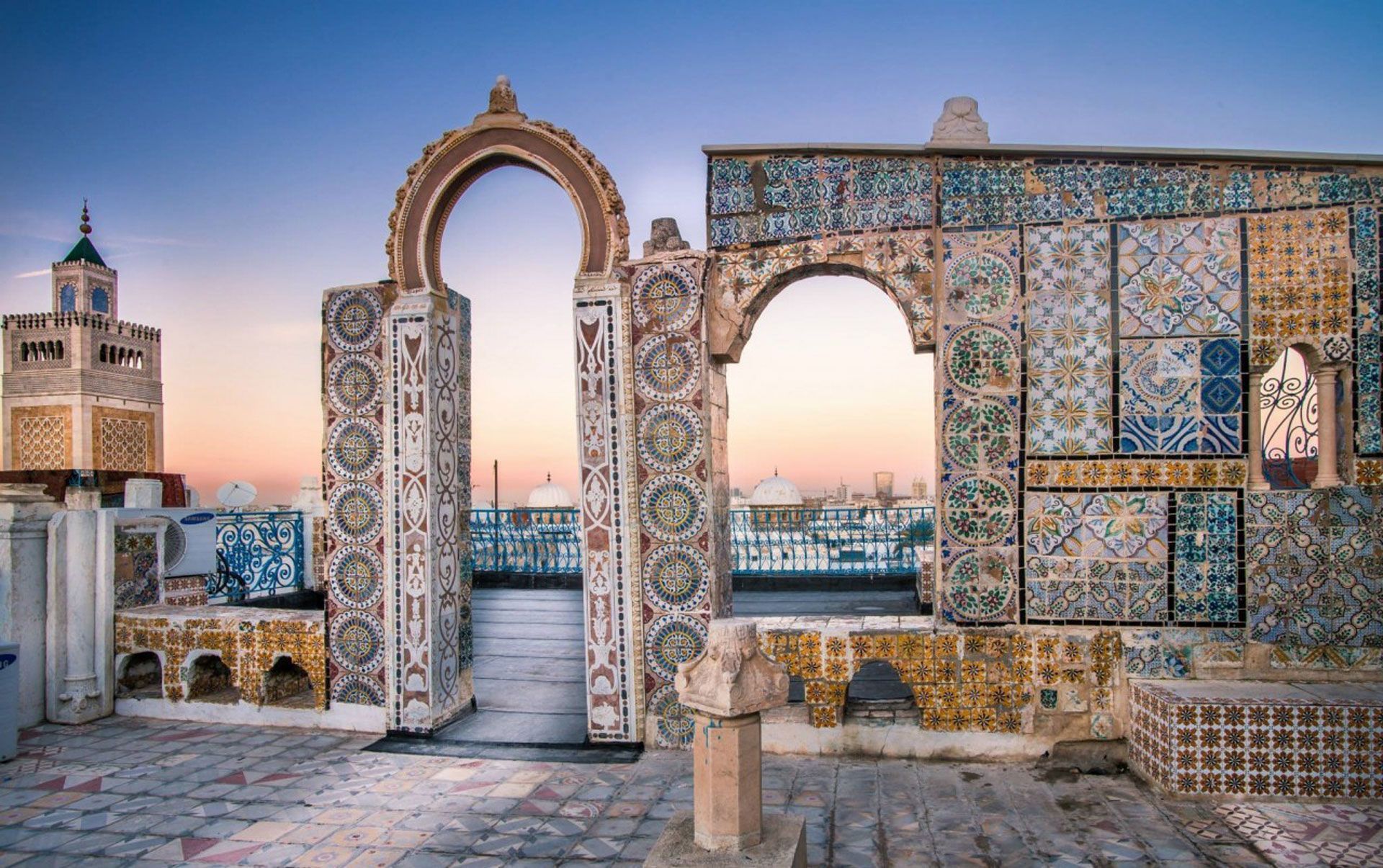 Une terrasse sur le toit avec des arches ornées et des mosaïques colorées, surplombant un paysage urbain avec un minaret au coucher du soleil.