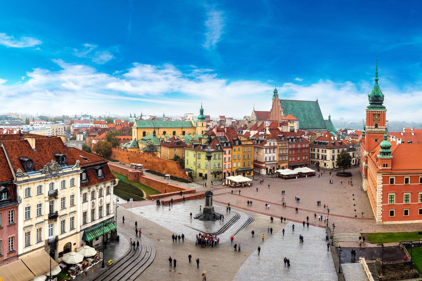 Una vista elevada de una plaza histórica europea llena de gente, rodeada de edificios coloridos y catedrales bajo un cielo azul.