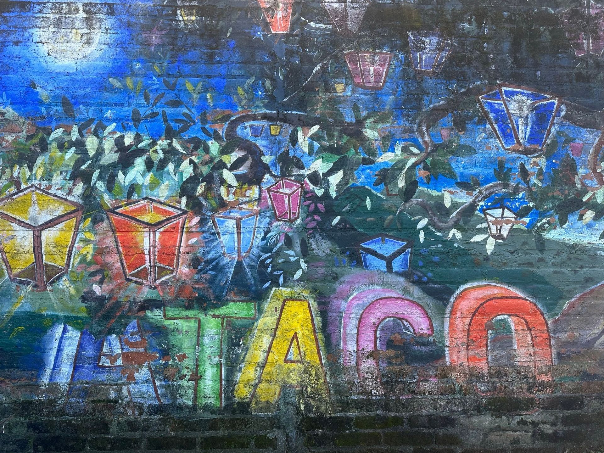 Ein farbenfrohes Graffiti-Wandbild auf einer Ziegelmauer, das einen Baum mit leuchtenden Laternen an den Ästen unter einem Vollmond zeigt.