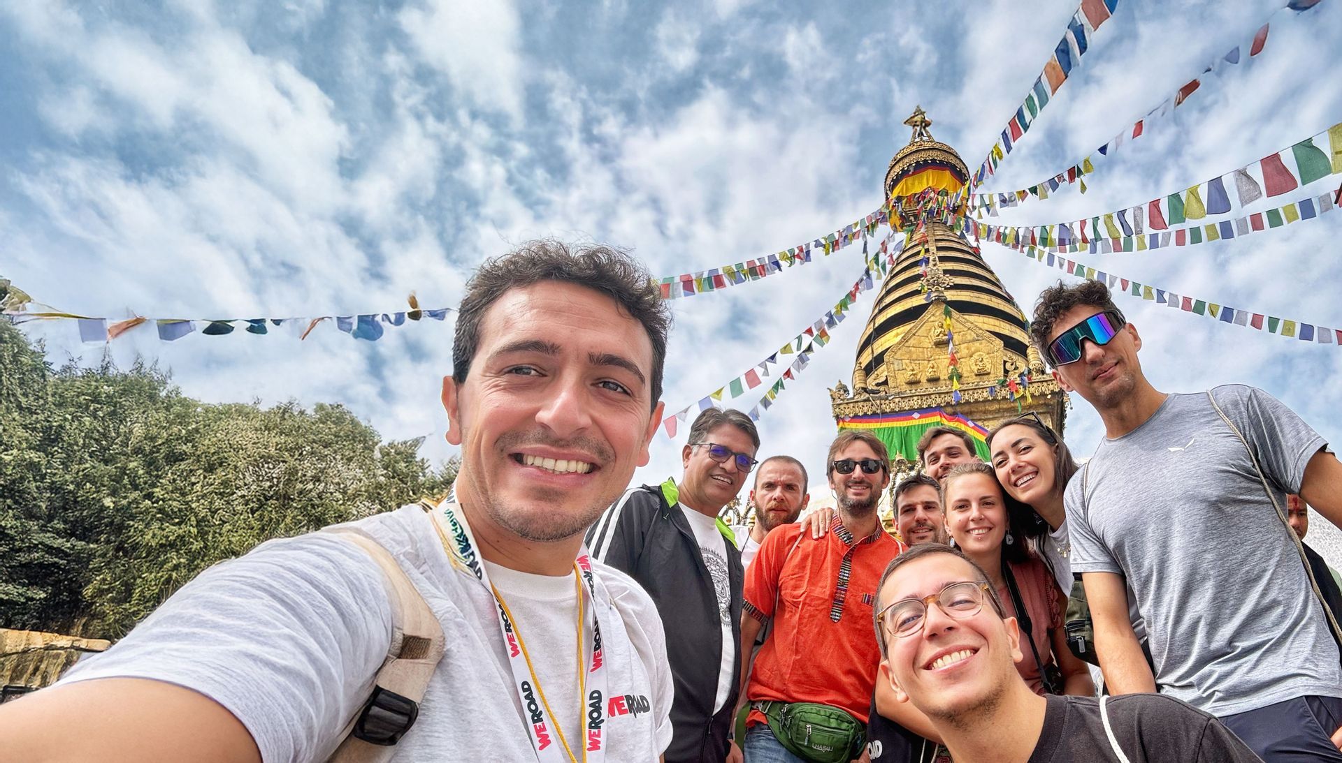Eine WeRoad-Gruppe macht ein lächelndes Selfie vor einer goldenen Stupa, mit bunten Gebetsfahnen, die vor einem bewölkten Himmel wehen.