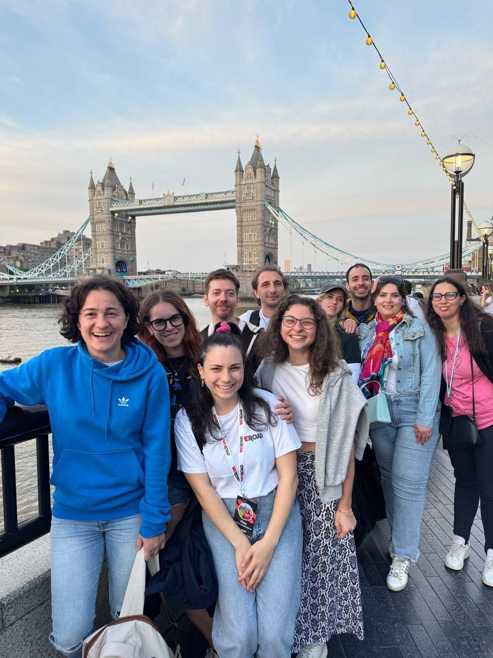 Un gruppo WeRoad in viaggio posa sorridendo per una foto su un lungomare, con un grande ponte storico visibile oltre il fiume.