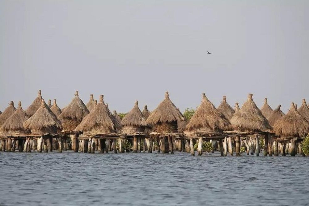 Ein Pfahlbautendorf mit konischen strohgedeckten Hütten steht in ruhigem Wasser unter einem dunstigen Himmel.
