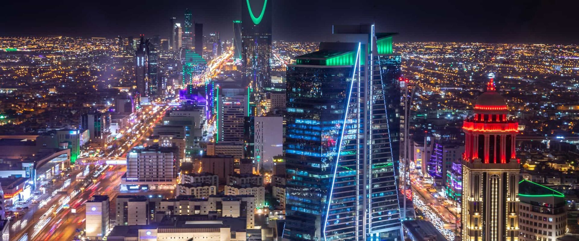 Un vaste panorama urbain nocturne, avec des gratte-ciel illuminés en vert, bleu et rouge, et les traînées lumineuses des véhicules dans les rues en contrebas.