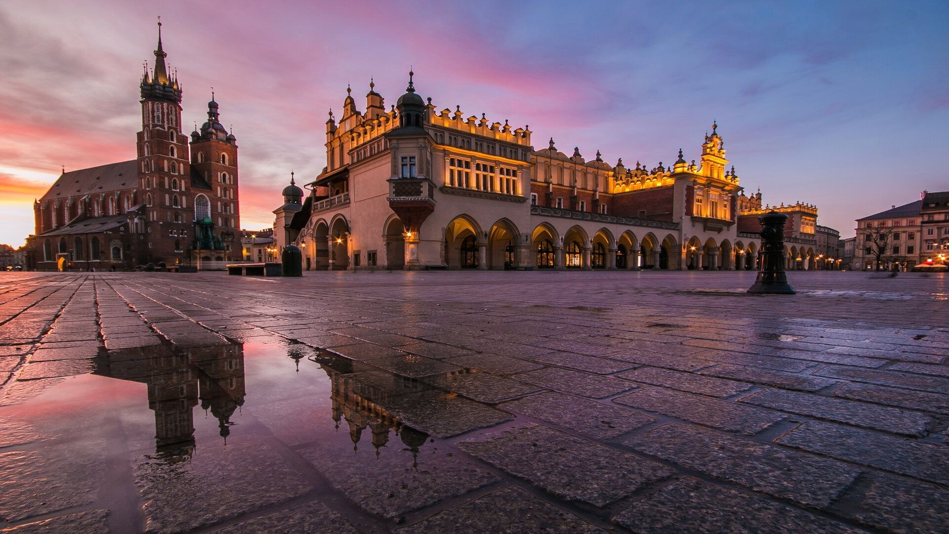 Una vista en ángulo bajo de edificios históricos iluminados en una plaza empedrada al atardecer, reflejados en un charco en el pavimento mojado.