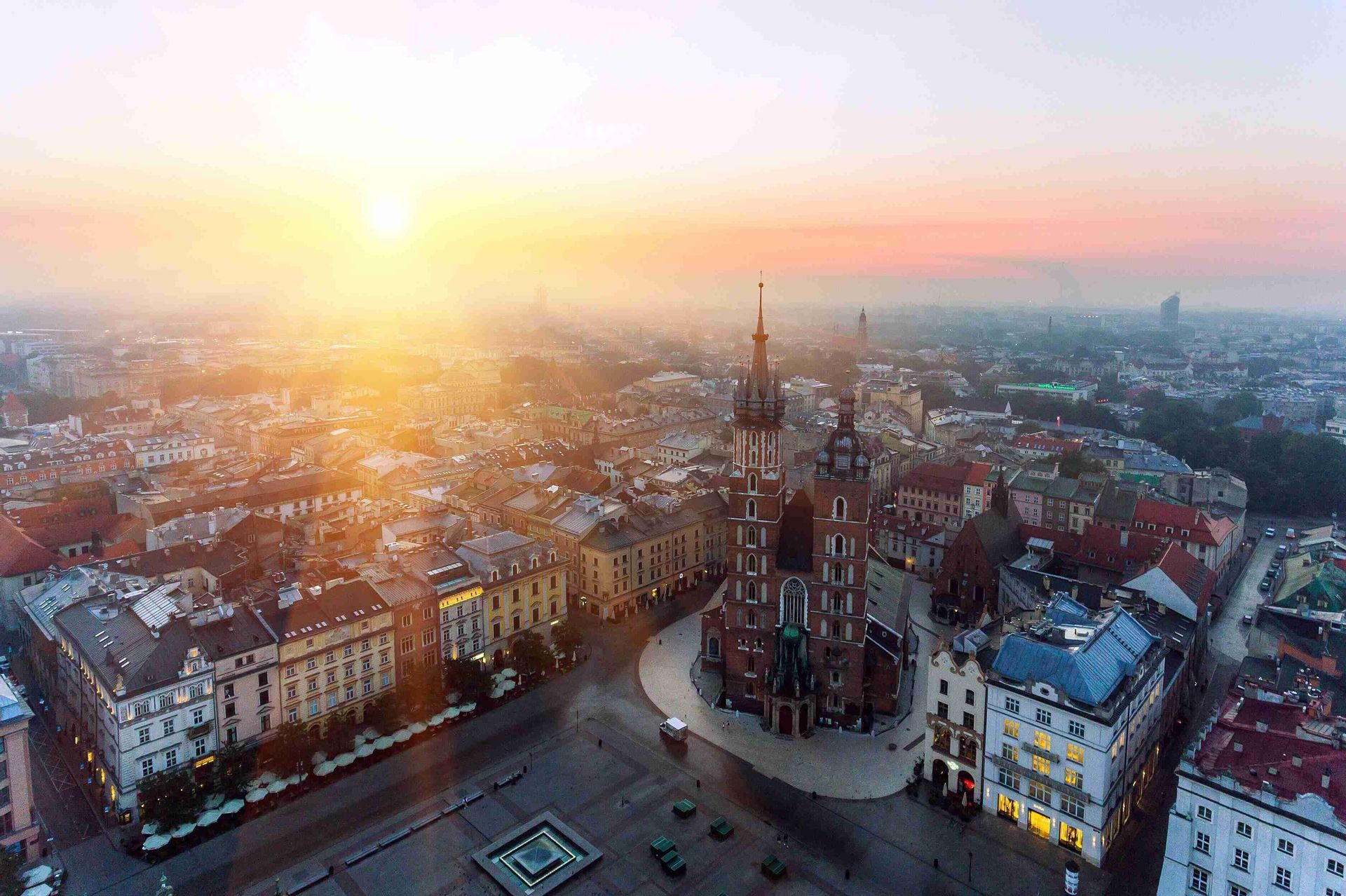 Una vista aérea de una plaza histórica y una gran iglesia de ladrillo bañadas por la luz dorada de un amanecer brumoso.